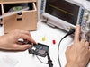 Hands testing an Arduino UNO R4 WiFi board connected to an oscilloscope, with a finger pressing a channel button on the oscilloscope and LEDs and components visible on the workbench.