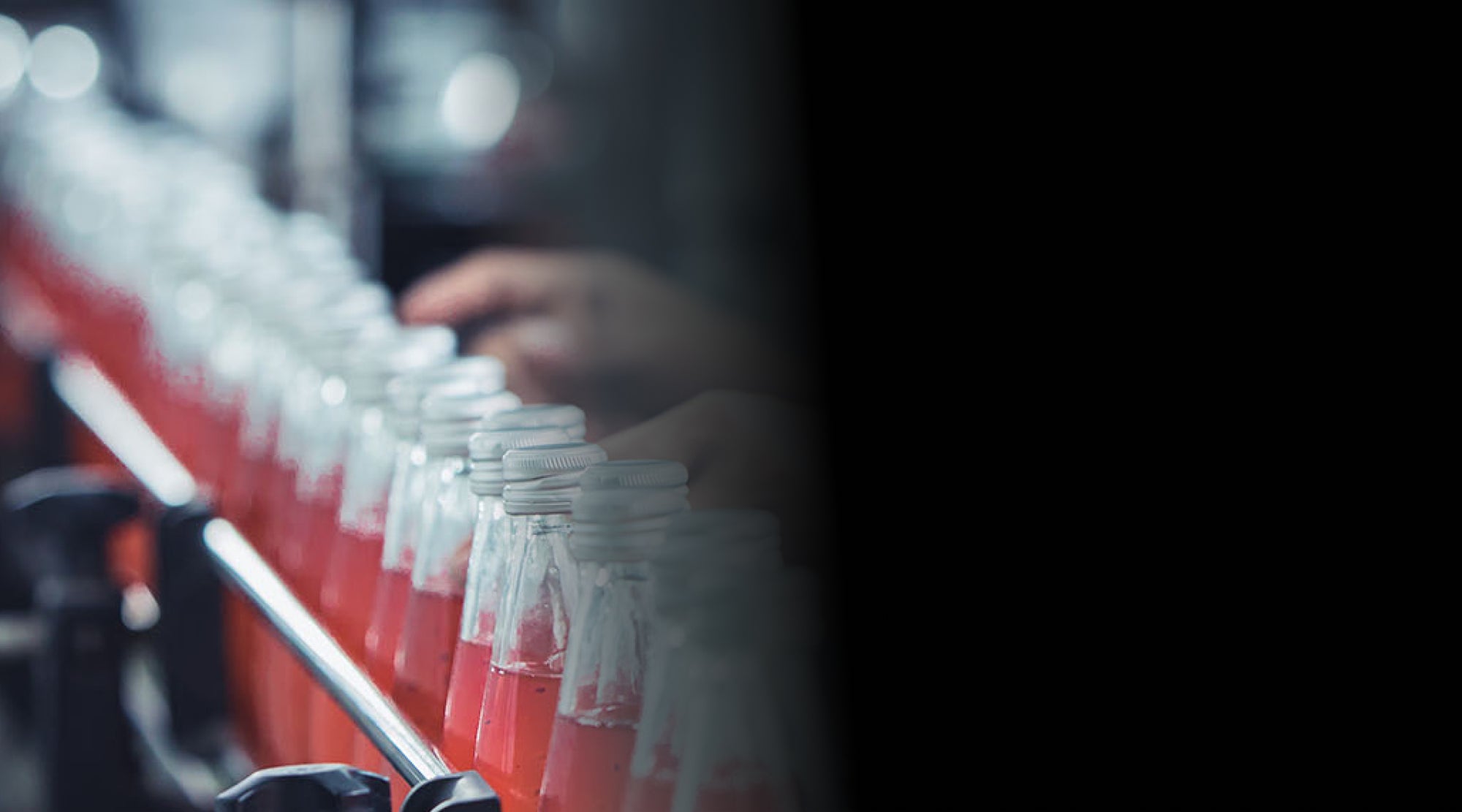Close-up of a production line with glass bottles filled with red liquid, sealed with caps, and blurred hands of a worker in the background.