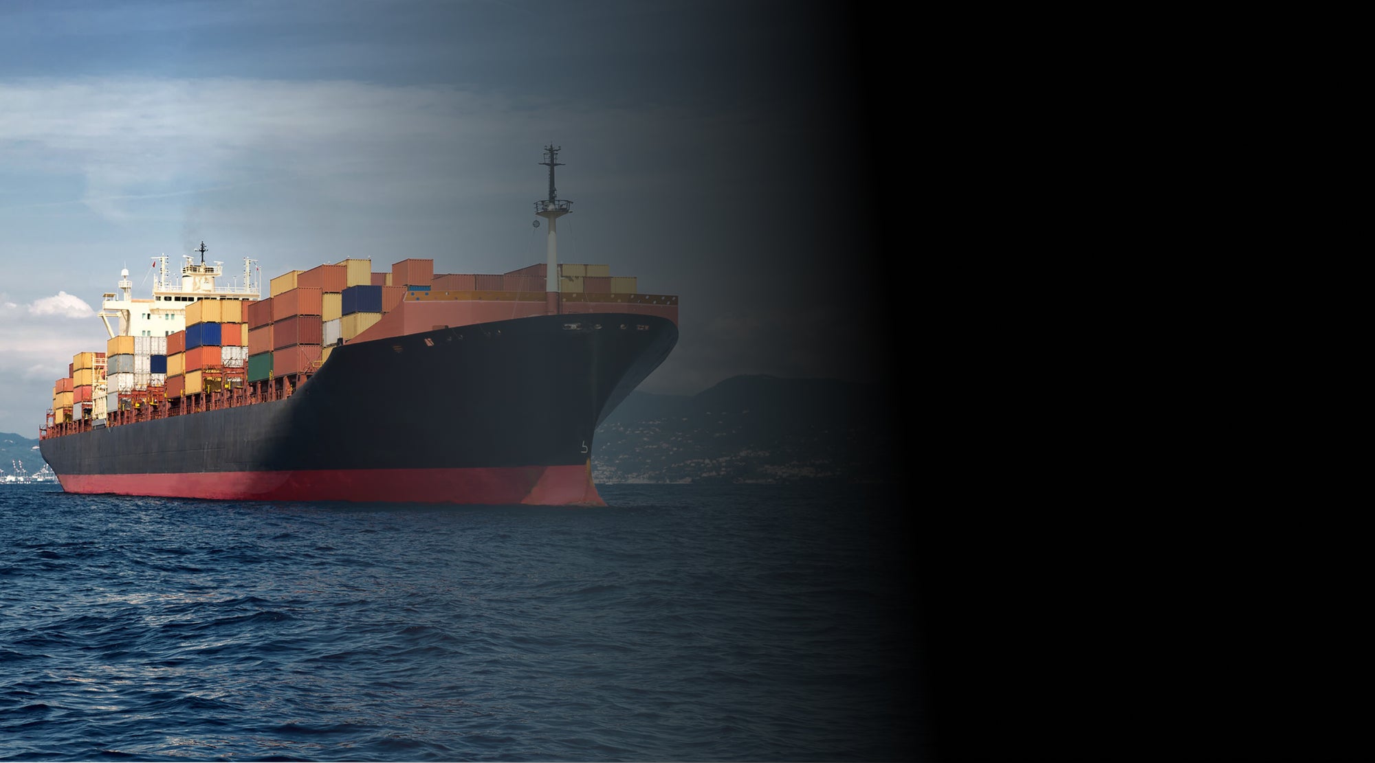 Large cargo ship loaded with colorful shipping containers navigating through open sea, with a coastal city and mountains visible in the background.